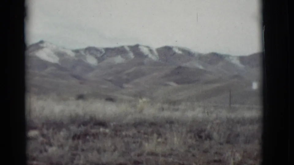 1964 home-movie frame showing a grassy New Mexico plain with snow-patched mountains under a pale sky.