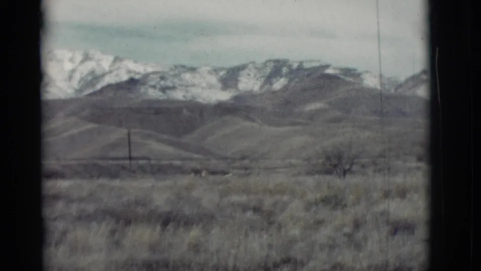 Distant snow-covered mountains and green-gray hills appear beyond a dry New Mexico foreground in this 1964 8mm frame.