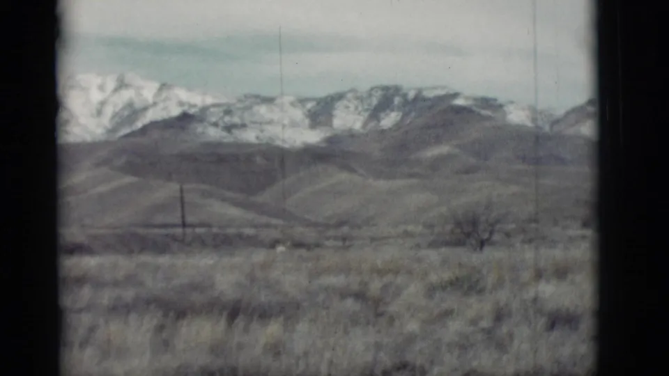 New Mexico home-movie frame showing dry grassland, a lone utility pole, rolling hills, and distant snow-covered mountains.