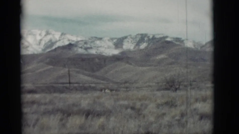 1964 8mm view of a bare shrub, dry grass, low New Mexico hills, and a distant snow-covered mountain range.