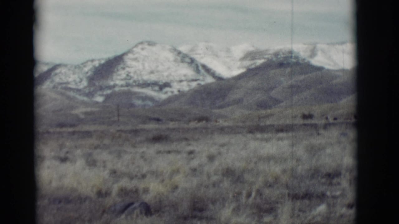 1964 New Mexico 8mm view of dry grassland leading to snow-patched mountain ridges under a pale sky.