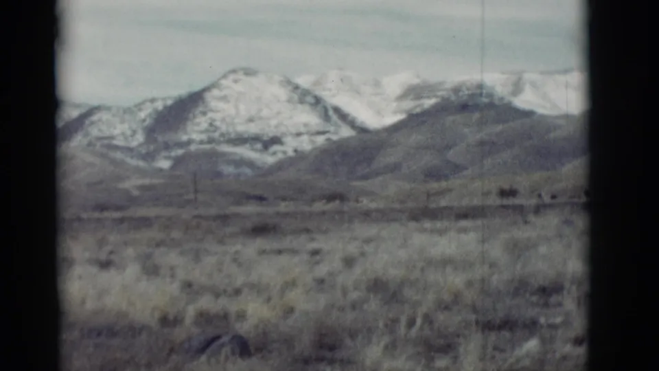 1964 New Mexico 8mm view of dry grassland leading to snow-patched mountain ridges under a pale sky.