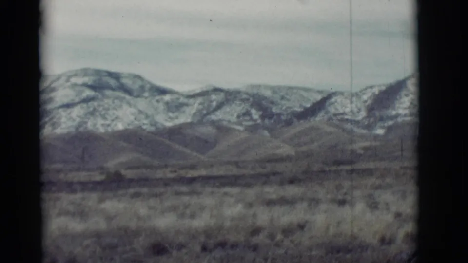 1964 New Mexico home-movie frame showing dry foreground grass, rolling hills, and distant snow-dusted mountains beneath an overcast sky.