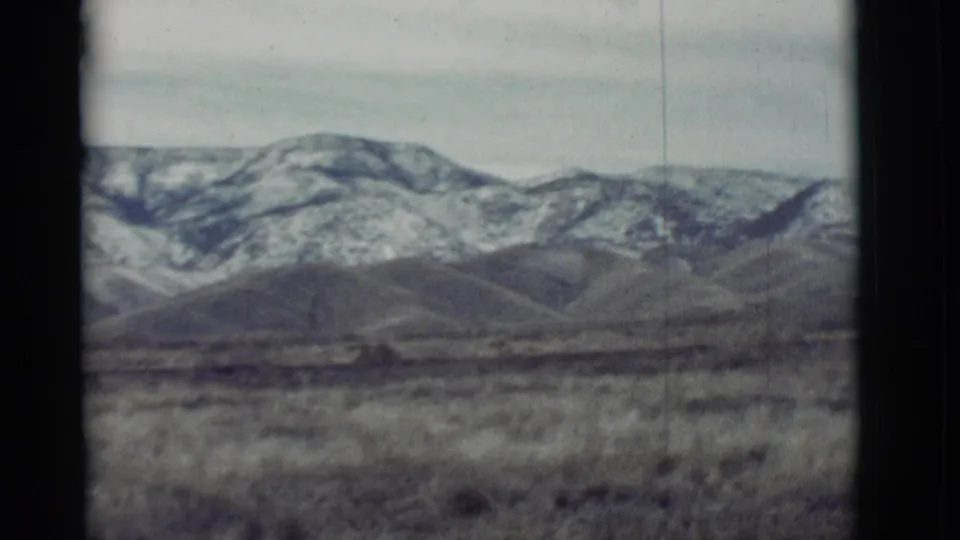 1964 New Mexico landscape frame with muted 8mm color, showing distant snowy mountains behind low hills and open dry terrain.