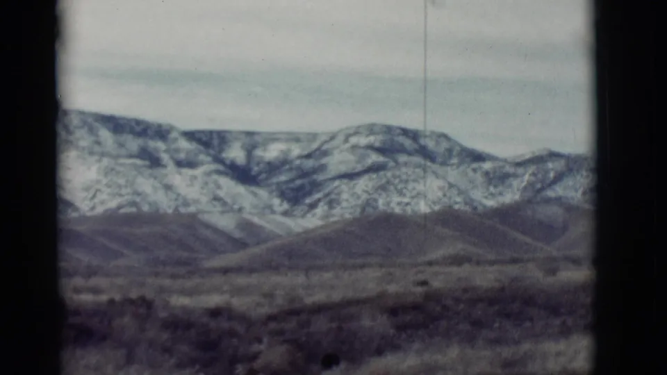 1964 New Mexico 8mm frame showing a closer view of snow-marked mountains, rounded foothills, and dry open ground.
