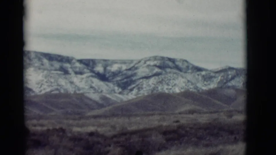 1964 New Mexico home-movie frame of open desert grassland, low hills, and a distant snow-dusted mountain ridge.