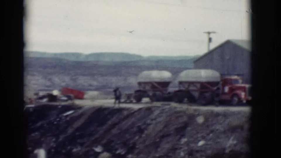 1964 New Mexico roadside yard with tank trailers, a red truck, a small walking figure, and mountains in the background.