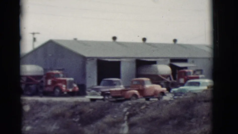 1964 New Mexico garage scene with classic cars, trucks, tank trailers, and a long utility building.