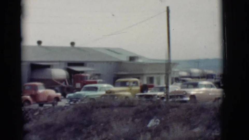 1964 New Mexico 8mm frame showing a line of classic cars and trucks outside a long garage near open terrain.
