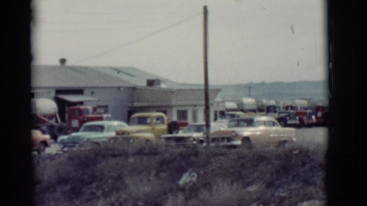 Classic cars and tanker trucks parked near a low roadside building.