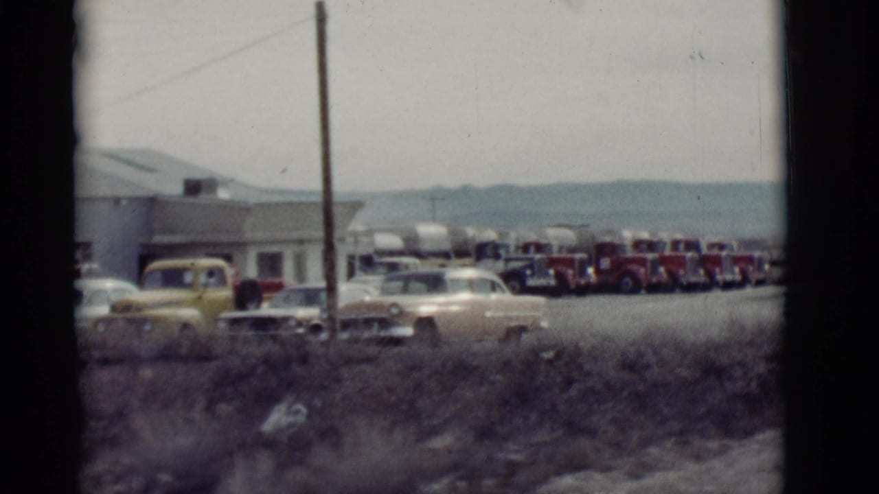 Parked sedans, a yellow pickup, and a line of tanker trucks near a roadside building.