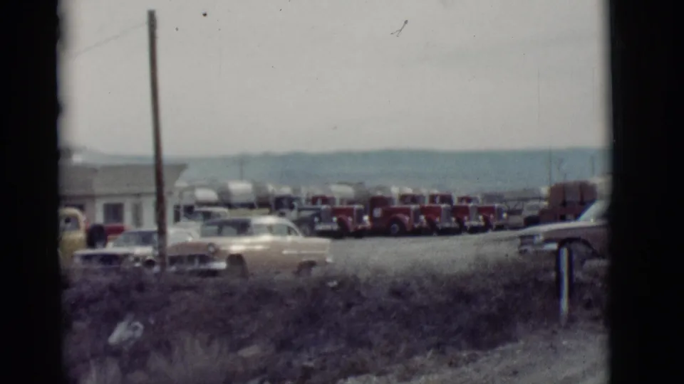 1964 New Mexico roadside scene with a pale sedan in front of tanker trucks, low structures, dry grass, and distant hills.