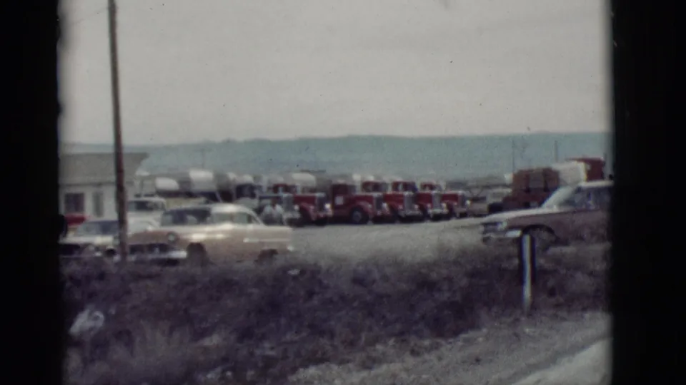 End frame of a 1964 New Mexico roadside stop with red tanker trucks, classic cars, a low building, pole, and dry foreground grass.