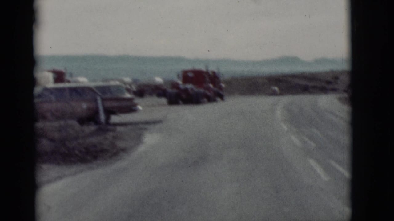 Curving paved road with parked cars and red trucks near open roadside terrain.
