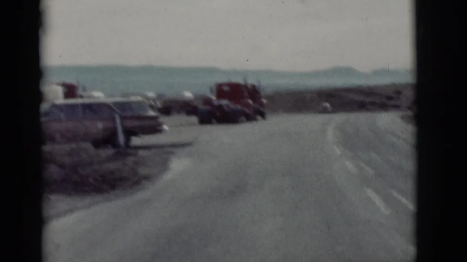 1964 New Mexico travel view looking along a curved paved road with parked vehicles and red trucks under a pale sky.