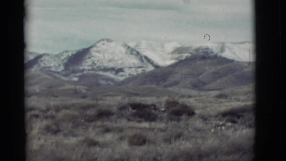 1964 New Mexico landscape frame showing scrubby desert foreground and snow-capped mountain ridges in soft 8mm focus.