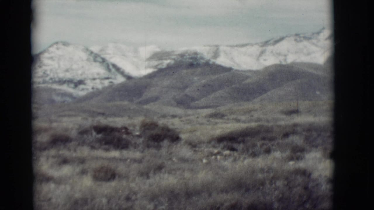 1964 New Mexico 8mm frame showing scrubby foreground hills and distant snow-dusted mountain peaks under a pale sky.