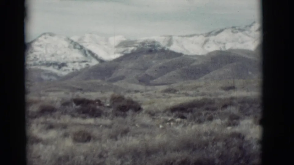 1964 New Mexico 8mm frame showing scrubby foreground hills and distant snow-dusted mountain peaks under a pale sky.