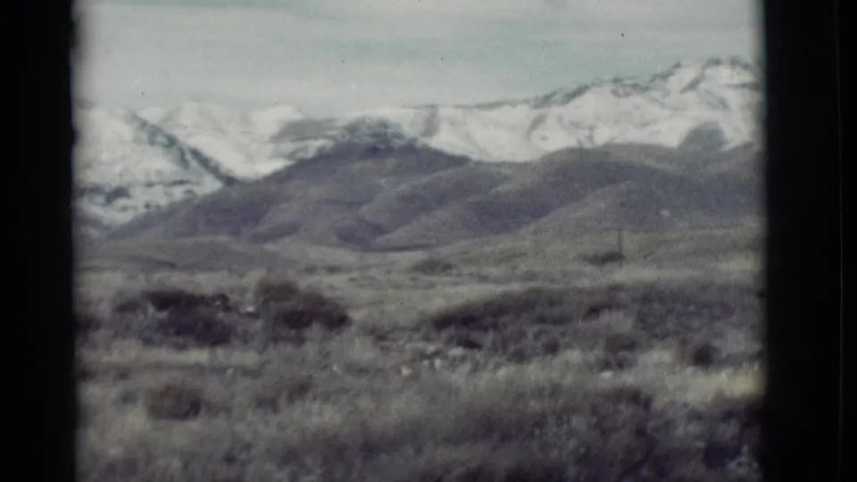 1964 New Mexico home-movie frame with muted 8mm color, dry brush, low foothills, and snow-marked mountain ridges.