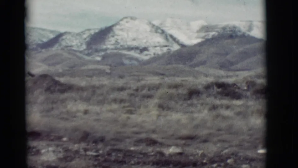 1964 New Mexico 8mm frame looking across dry ground toward snow-patched mountains and darker foothill slopes.