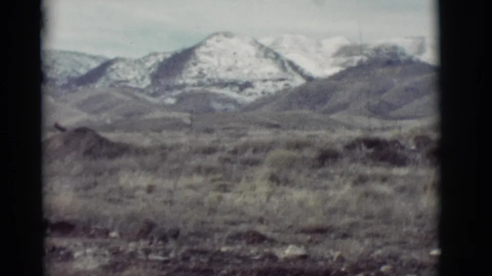 1964 New Mexico home-movie frame showing a snowy mountain peak, dark foothills, and dry open land in soft 8mm focus.