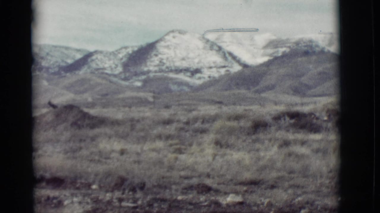 1964 New Mexico 8mm frame showing snowy mountain peaks across an arid scrub foreground under a pale sky.