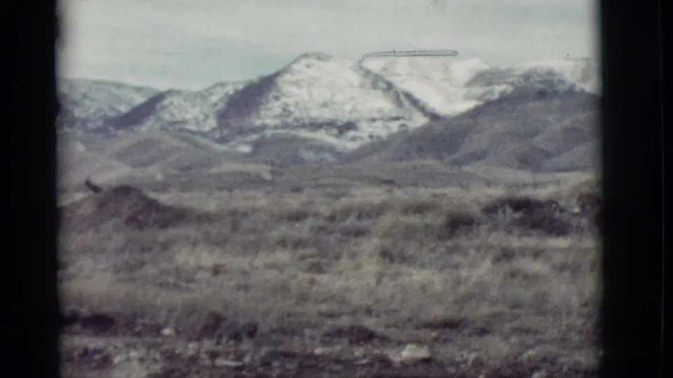1964 New Mexico 8mm frame showing snowy mountain peaks across an arid scrub foreground under a pale sky.