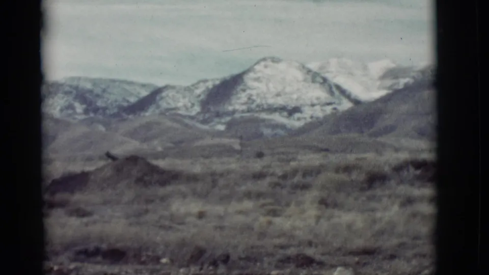 1964 New Mexico 8mm frame showing arid foreground brush, rolling hills, and distant snow-capped mountains.