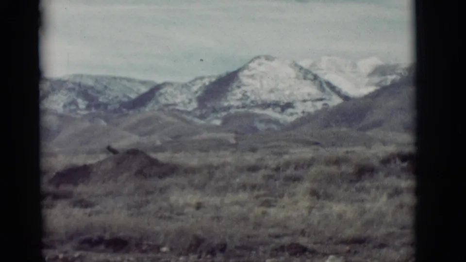 1964 New Mexico landscape frame with sparse desert vegetation, rounded foothills, and snow-covered mountain peaks.