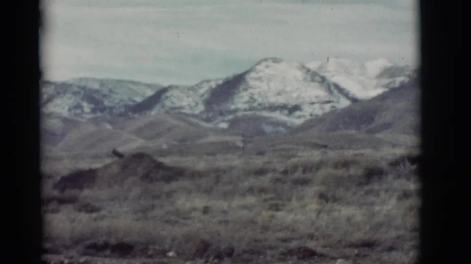 New Mexico home-movie frame from 1964 showing scrubby desert terrain below distant snow-capped mountains.