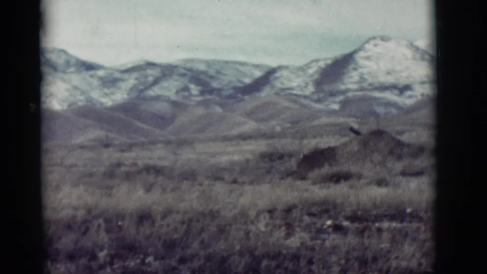1964 New Mexico 8mm frame showing dry foreground grasses, rounded hills, and distant snow-covered mountains under a pale sky.