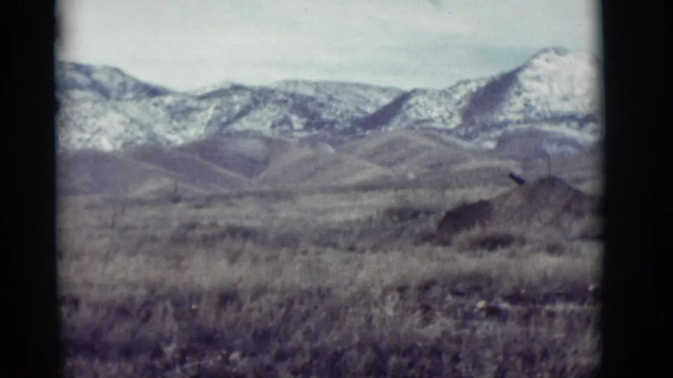 New Mexico landscape frame from 1964 with snow-dusted mountains, low foothills, dry grasses, and dark 8mm frame edges.