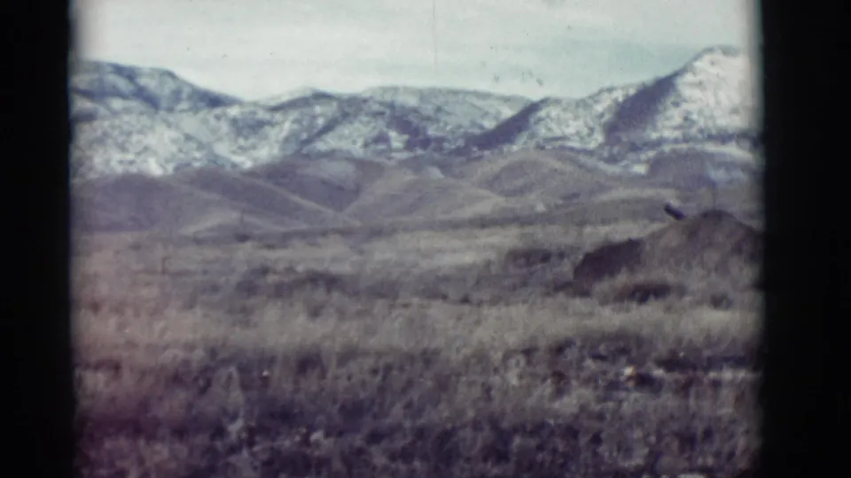 1964 New Mexico 8mm frame showing dry foreground grass, rolling hills, and snow-dusted mountains in the distance.