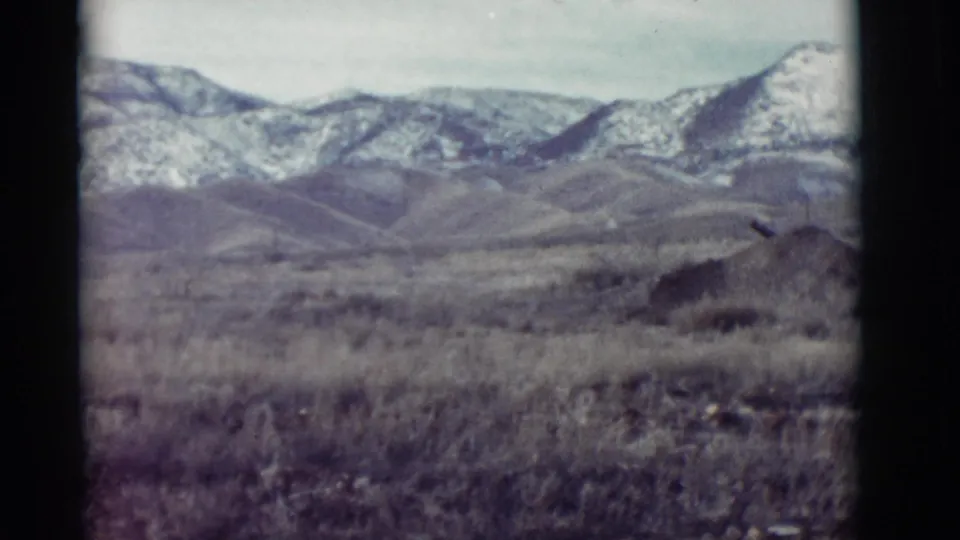 1964 New Mexico home-movie view of sparse grassland, rounded hills, and a distant snow-marked mountain ridge.