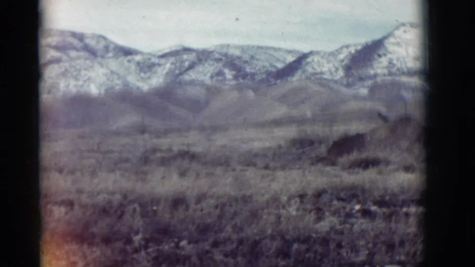 Soft 1964 New Mexico 8mm frame with dry vegetation in front of rolling hills and snow-dusted mountains.