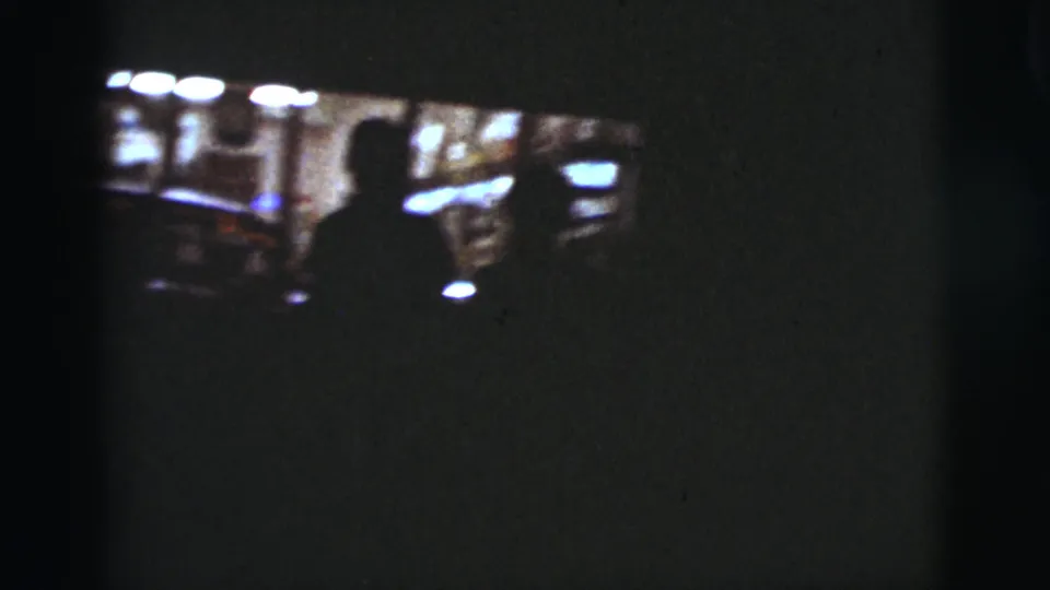 A dim 1960s home-movie frame from inside a vehicle, with a silhouetted person near bright car windows.