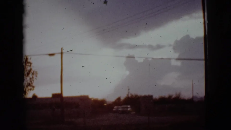 A classic car drives along a low suburban road in Arizona beneath heavy storm clouds and power lines.