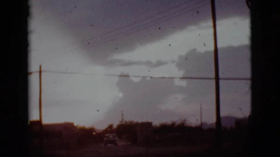 1965 Tuscon, Arizona 8mm frame showing storm clouds above a quiet road, utility lines, and a small car in the distance.