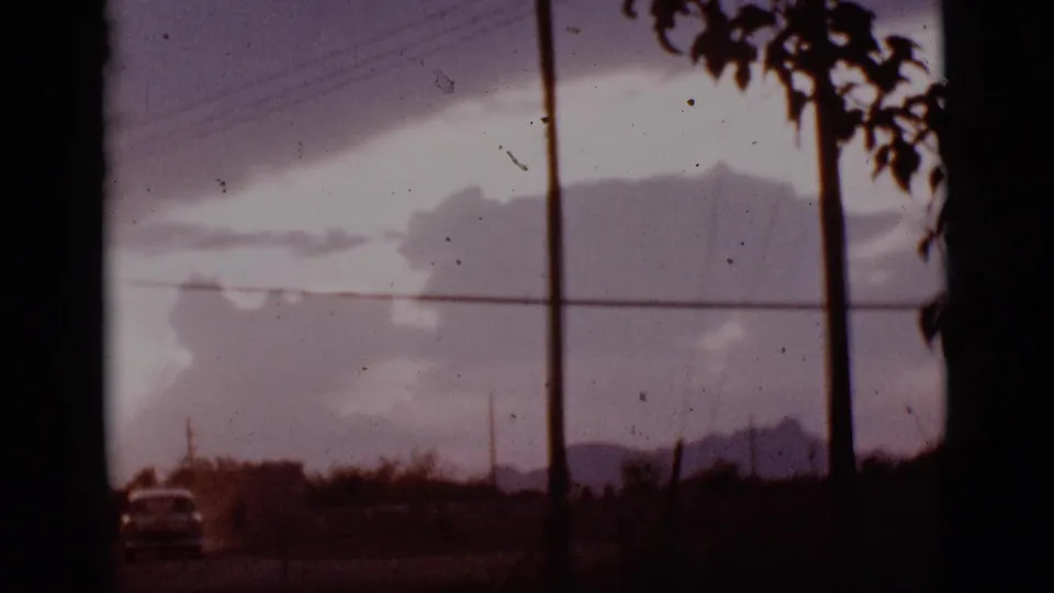 1965 Tuscon, Arizona 8mm frame with heavy clouds, crossing power lines, a distant car, and foliage entering from the upper right.