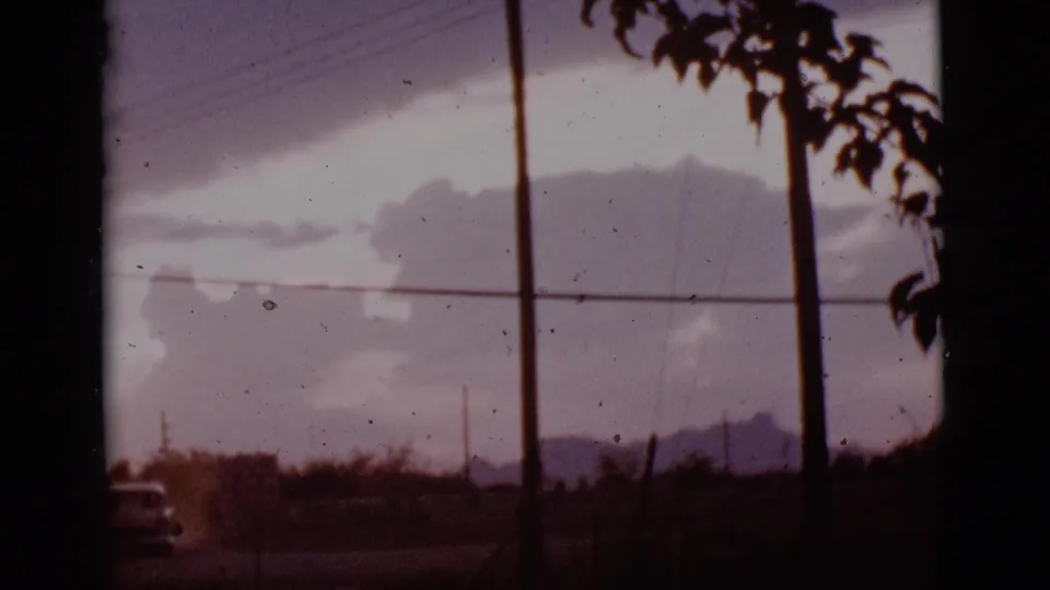 1965 Tucson-area 8mm frame with distant mountains, clouds, utility poles, wires, and dark tree silhouettes from a car-view perspective.