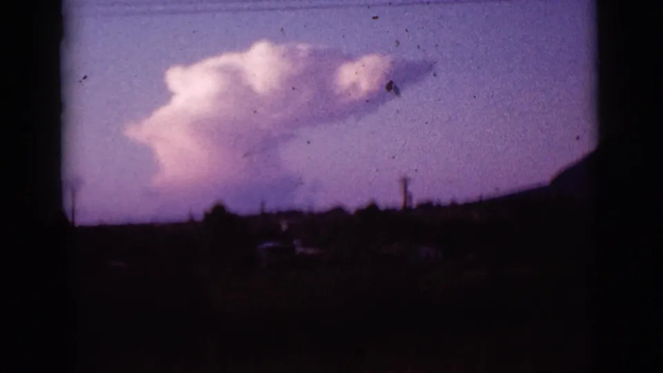 1965 Tucson-area 8mm frame with a large billowing cloud above a dark mountain ridge and faint roadside forms near the lower edge.