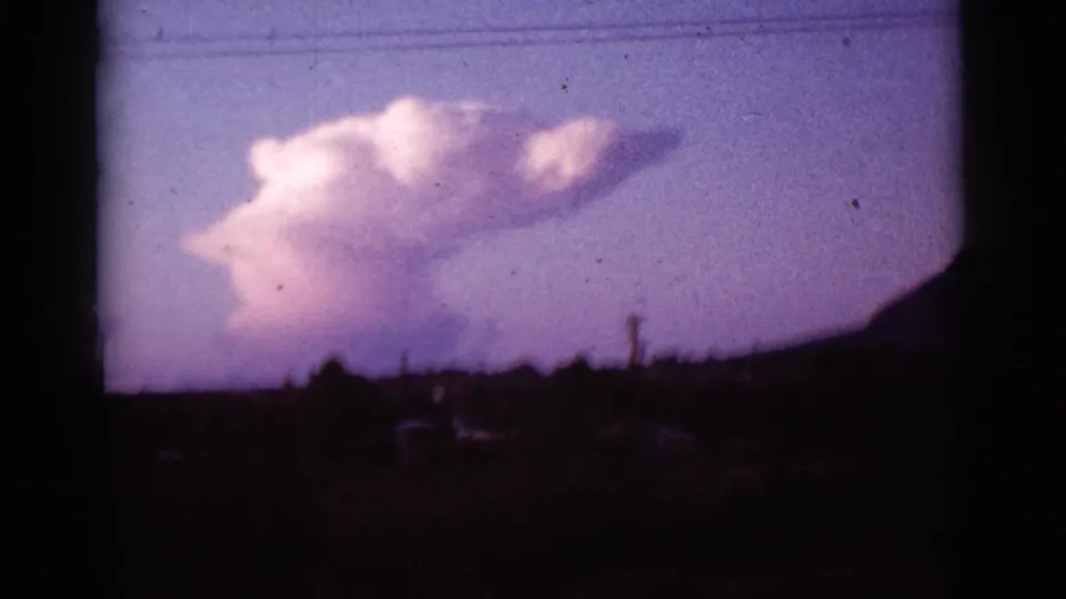 1965 Tucson-area 8mm frame showing a bright cloud, silhouetted mountain ridge, overhead wires, and dark roadside foreground.