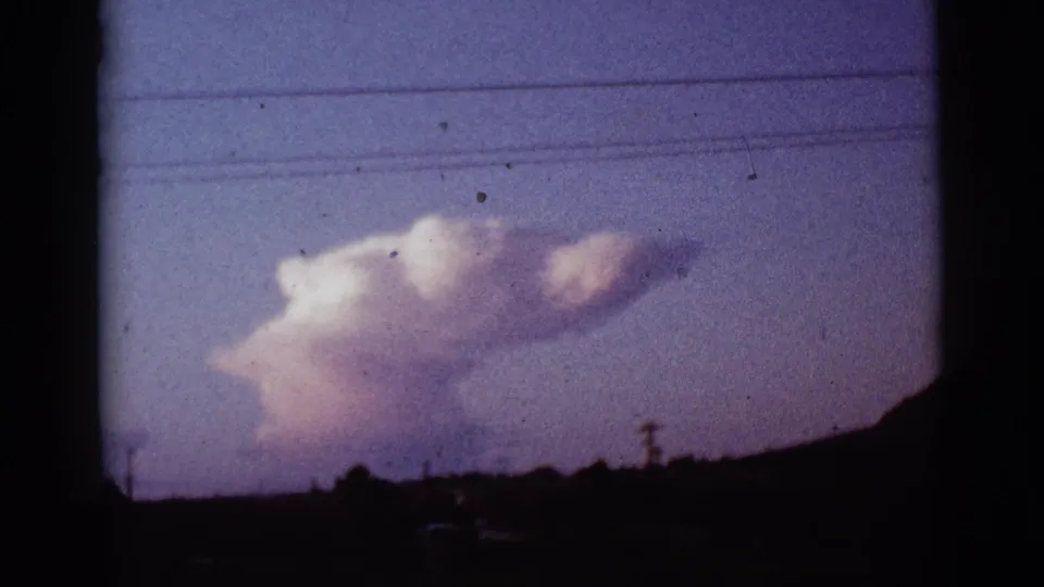 1965 Tucson, Arizona home-movie frame with a pale pink-white smoke plume above a dark horizon and power lines.