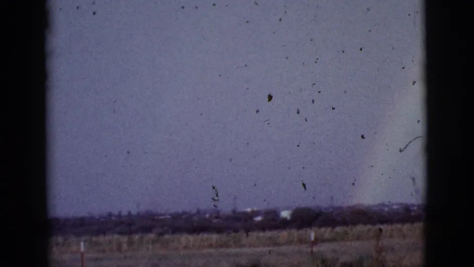 1965 Tuscon, Arizona 8mm frame showing a faint rainbow rising over scrubland, with fence posts and distant vegetation below a washed sky.