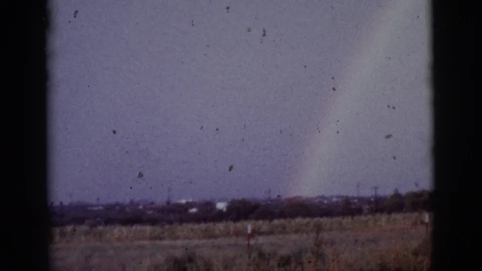 1965 Tuscon, Arizona 8mm frame showing a faint rainbow on the right side of a purple-tinted sky above scrubland and a low distant horizon.
