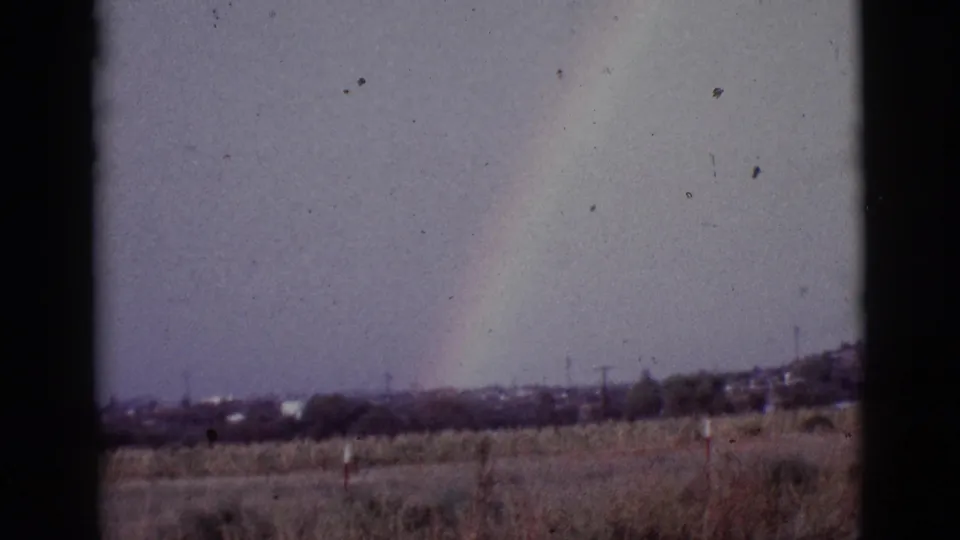 1965 8mm frame showing a soft rainbow over a rural Tucson-area horizon with utility poles, brush, and film grain.