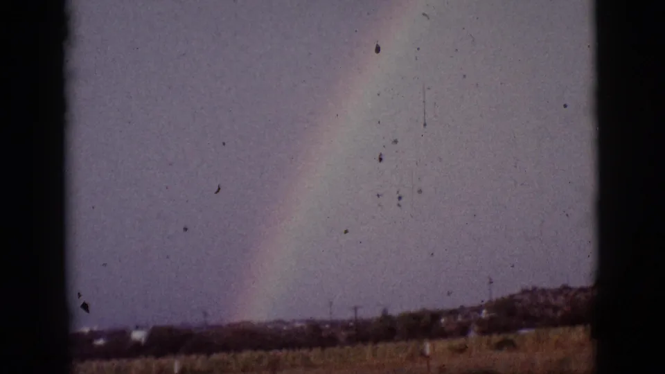 1965 8mm home-movie frame with a pale rainbow over dry Arizona grassland and a low distant hillside.