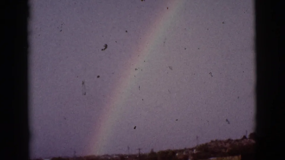 1965 8mm frame of a rainbow band over low Tucson-area brush, with soft color, grain, and speckling from the film scan.