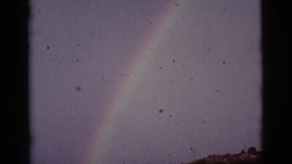 1965 8mm Tucson-area home-movie frame showing a faint rainbow against a muted sky above a low, indistinct horizon.
