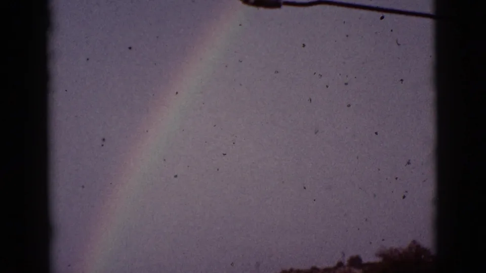 1965 8mm Tucson-area home-movie frame showing the rainbow dropping toward distant ground, with a dark vertical edge and wire in view.
